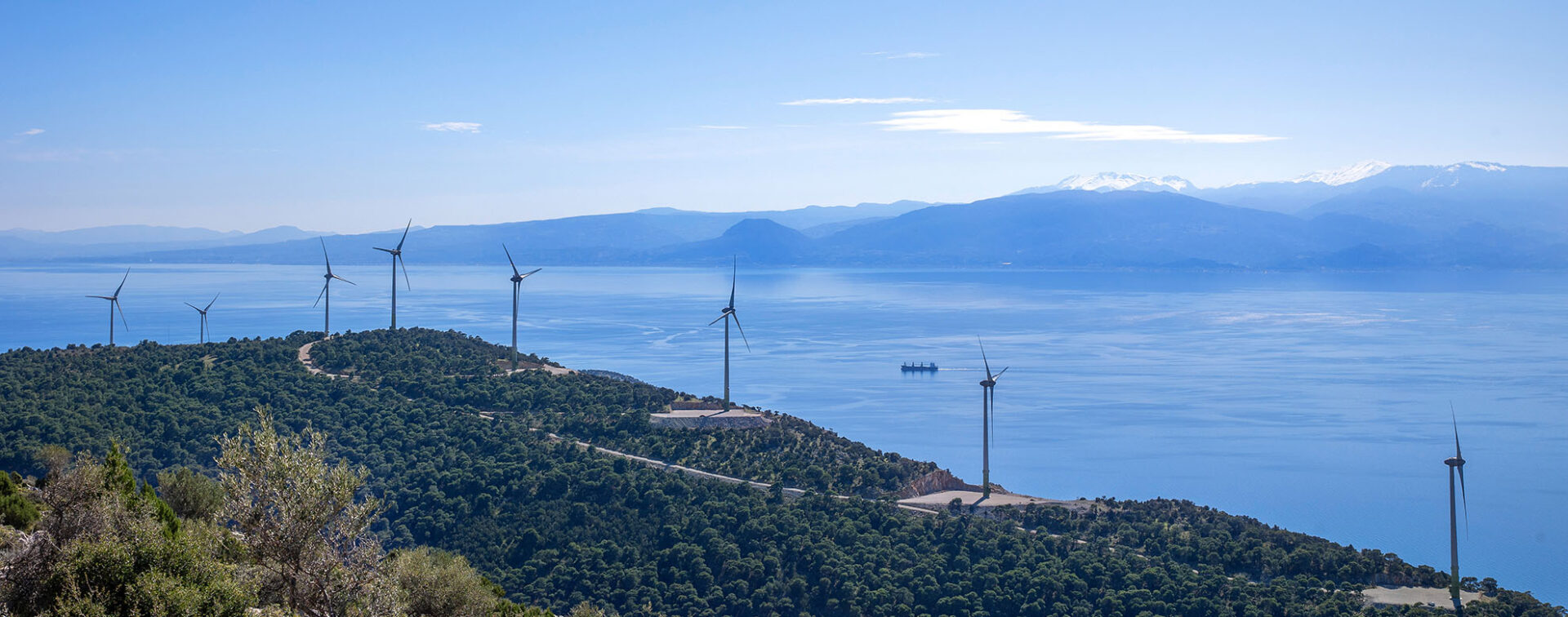 Windfarm with 8 windmills on green hills in front of the sea with hazy mountains in the back and blue sky.