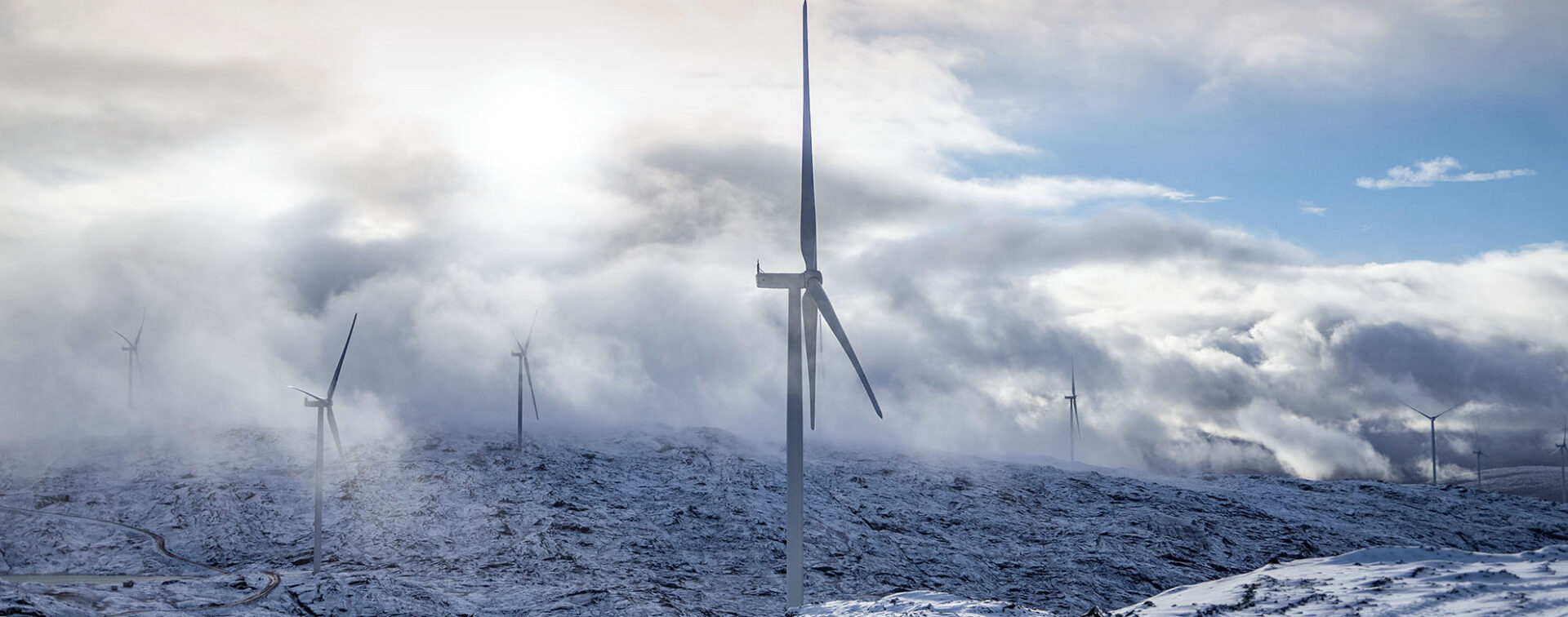 Windfarm on rocky hills with 8 windmills, some barely visible in the mist of a heavily cloudy sky.