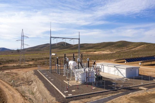 Fenced battery storage facility in sandy area with green bush-covered hills in background.