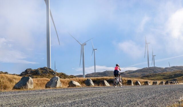 Woman cycling on gravel path with hills and windmills in background.