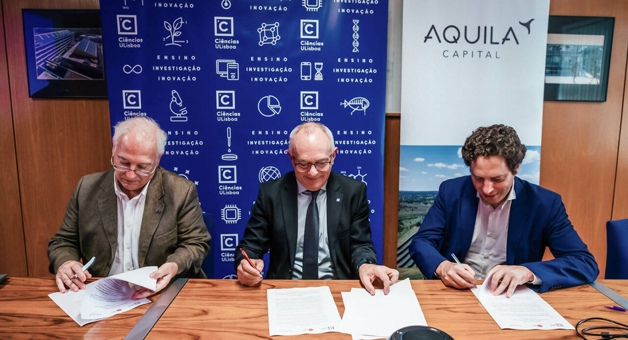 Three smiling middle-aged men in suits sit at a table in an office room and sign a contract.