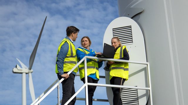 Three engineers wearing reflective vest in front of a windmill entry.