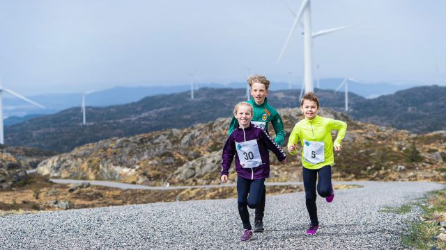 Three kids running and playing in the hills and windmills in the back.