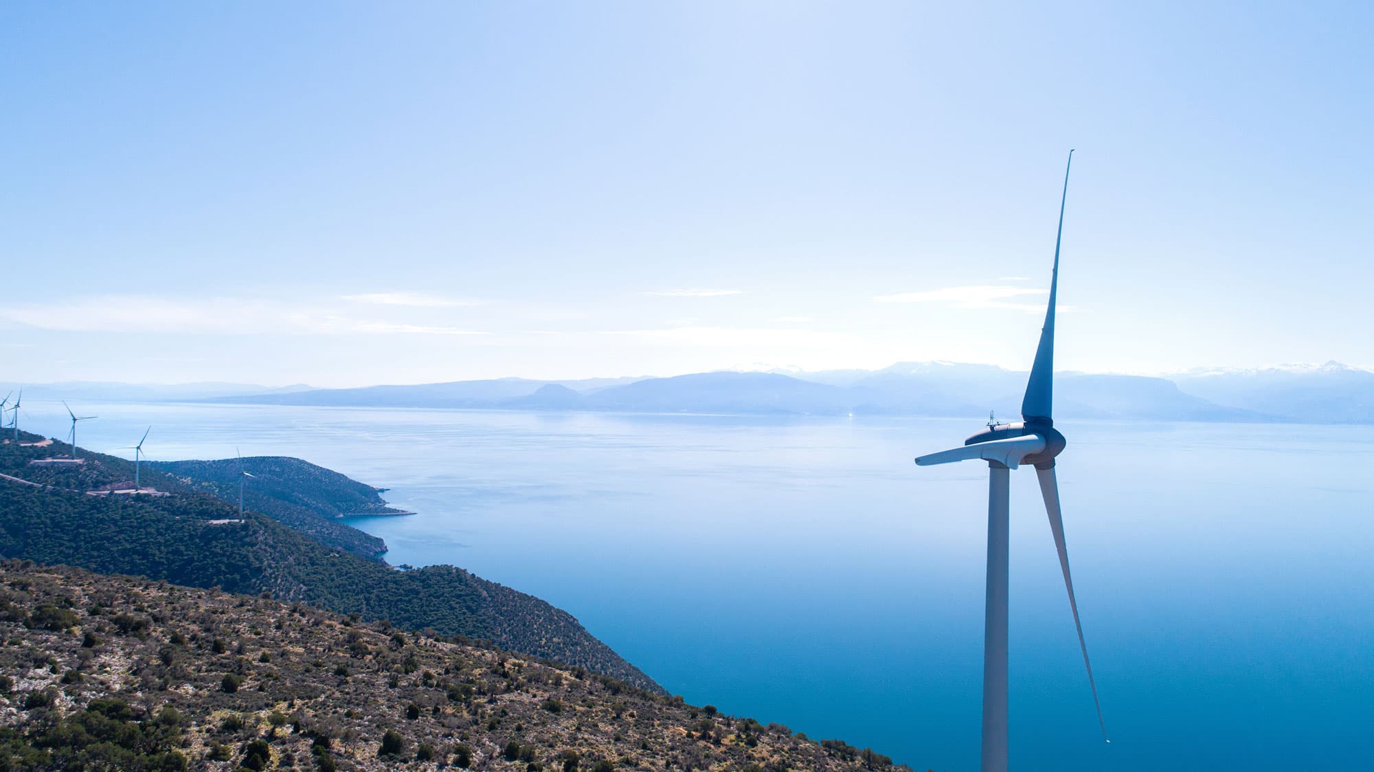 Windmills on green hills in front of water with mountains in the back and blue sky.