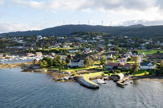 Small town on the shore with green hills and bigger mountains and wind turbines in the background.