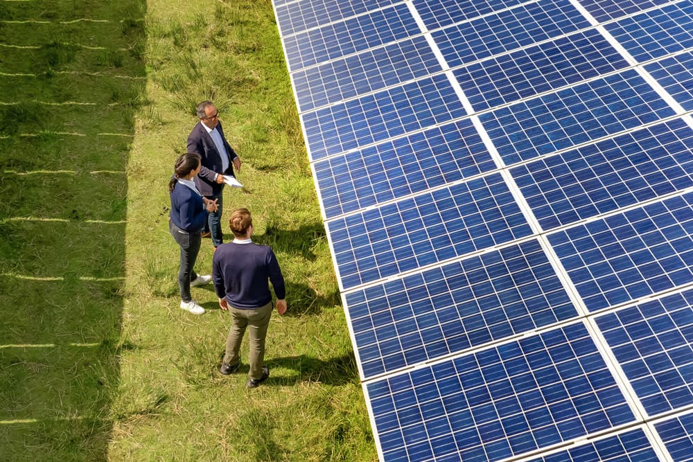 Three people in business clothes are standing in a solar park on a green grass field and are talking to each other.