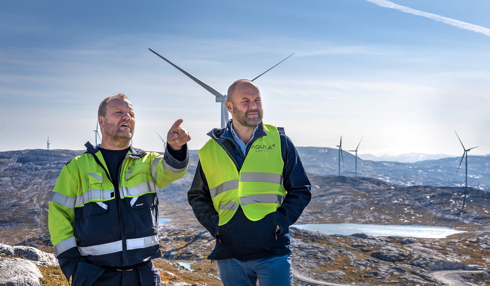 Two men wearing reflective vest and talking in front of windmill plant in hilly landscape.