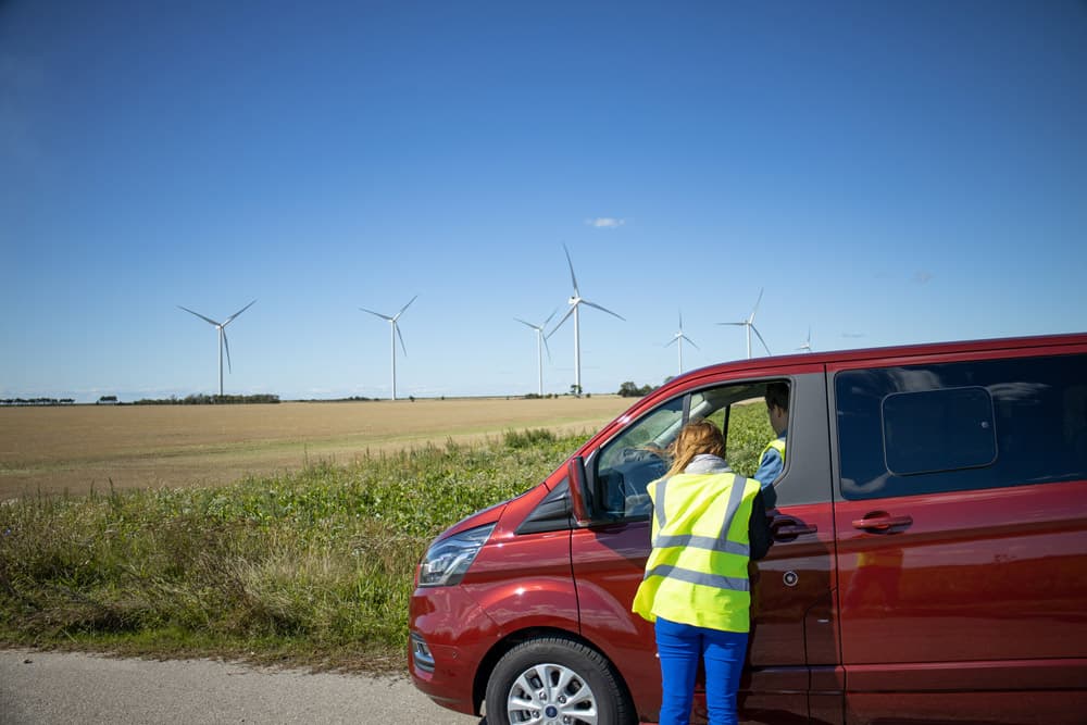Woman in yellow reflective vest talking to a van driver with fields and wind turbines in the background